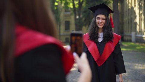 Cheerful graduate woman throwing mortarboard cap as friend taking photo on smartphone. Portrait of happy excited Caucasian student rejoicing success at university campus outdoors