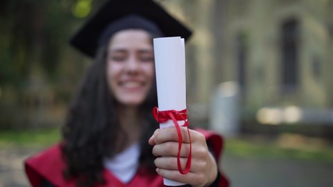 Close-up diploma with red ribbon in female hand with blurred smiling young woman at background. Happy Caucasian intelligent student in graduation toga and mortarboard cap at university campus