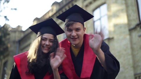 Joyful couple in graduation togas and mortarboard caps waving smiling looking at camera. Positive Caucasian boyfriend and girlfriend posing at university campus celebrating success outdoors