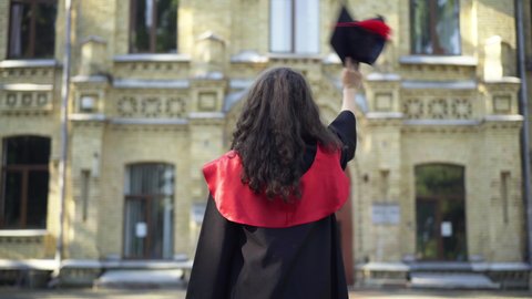 Back view slim young woman in graduation toga waving goodbye with mortarboard cap to university campus. Happy Caucasian graduate student outdoors on college yard in the morning