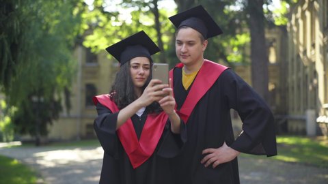 Satisfied couple of graduate students taking selfie on smartphone standing at university campus outdoors. Portrait of happy intelligent boyfriend and girlfriend in graduation togas enjoying morning