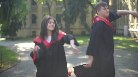 Satisfied excited graduates dancing on sunny morning at university campus smiling. Portrait of happy positive Caucasian groupmates in graduation togas having fun outdoors. Education and success