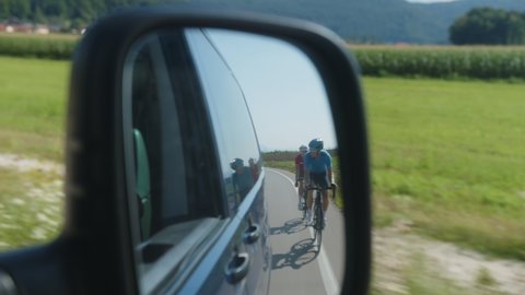 Two racing cyclists riding on a paved road, shot from the van, view in the rear view mirror.
