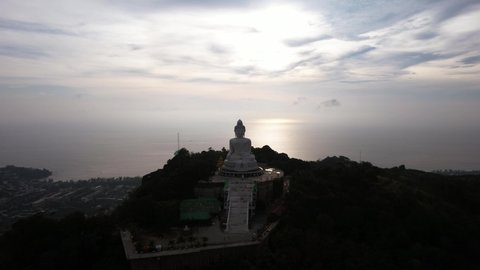 Drone view of the Big Buddha, Thailand. The Big Buddha is sitting on hill in the lotus position, meditating. The sun shines brightly through clouds. There's a jungle all around. A ray of sun on water