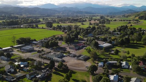 Aerial pull back of Tapawera township, rural area known for ho production, New Zealand. Green farmland, houses in valley, mountains in horizon.