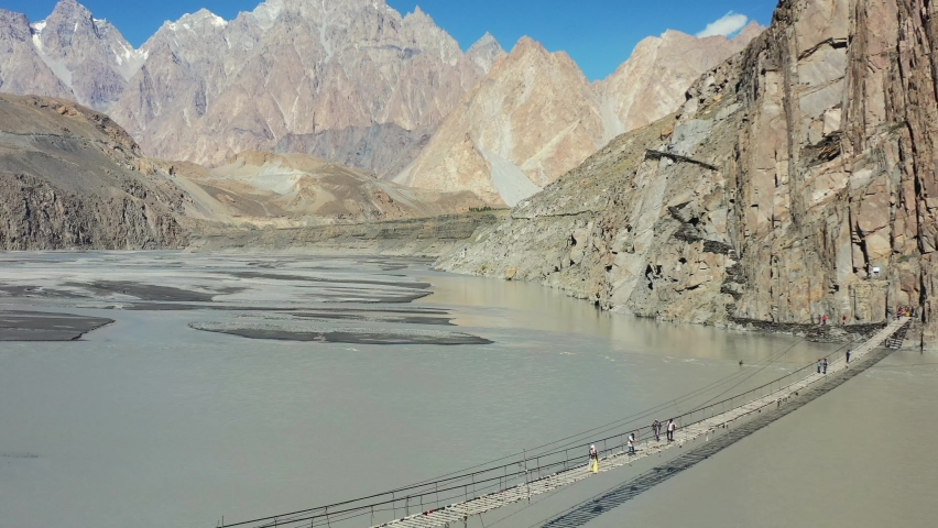 Aerial drone of tourists crossing the famous Hussaini bridge in Hunza Pakistan with a fast river flowing below and the Passu Cones mountains in the distance