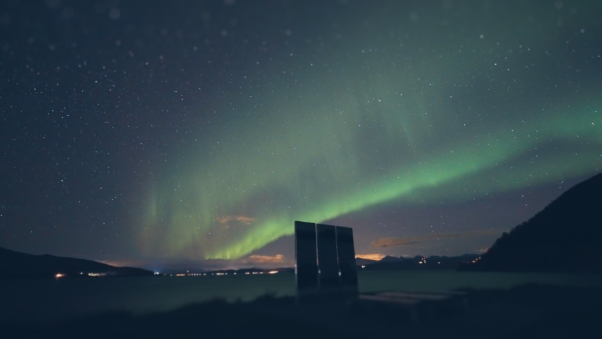 Aurora borealis above the Battle for Narvik memorial on the shore of the fjord. Street and car lights flash in the background. Timelapse.