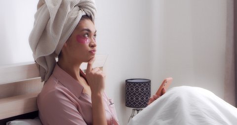 Young woman in satin pink pajamas with a towel on head and patches on her eyes Is sitting under a white blanket on bed in bedroom, planning a bachelorette party,making a to-do list on her cell phone