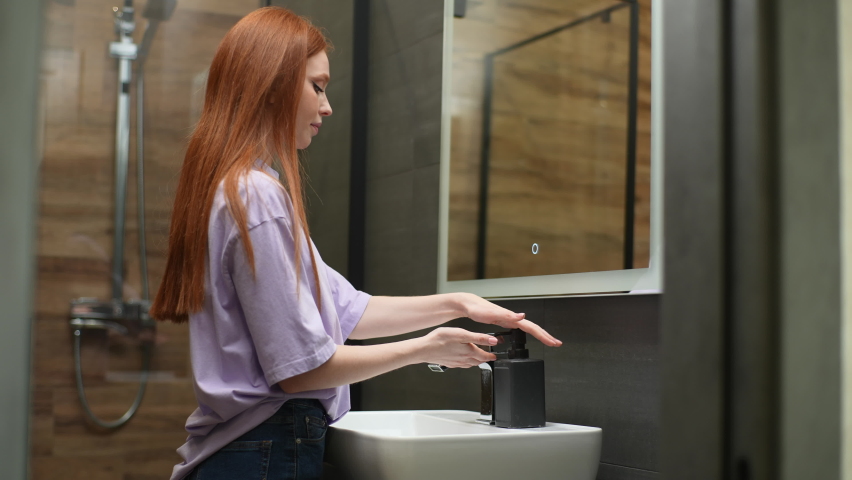 Side view of attractive young woman taking liquid soap and washing hands under tap water at home restroom with modern interior. Pretty female washing hands in sink after going to bathroom.