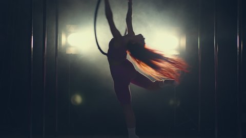 The silhouette of a girl approaches the gymnastic hoop against the background of a flashing light, takes hold of it with her hands and gracefully rotates, throwing back her head and periodically