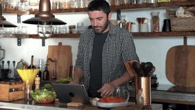 Handsome bearded man watching video recipe and preparing food at home in kitchen. Male cuts vegetables at the table for a vegetarian salad. healthy eating concept. 4k footage