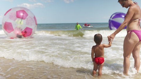 A little cute cheerful kid, looks at horizon of blue calm sea and bright inflatable big balls for children with joyful kind happy mother in bright bikini under the scorching sun. 4K UHD slow-mo video