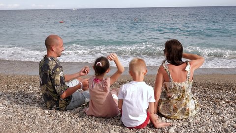 A cheerful family with little children are sitting on the seashore, they throw pebbles into the water together.