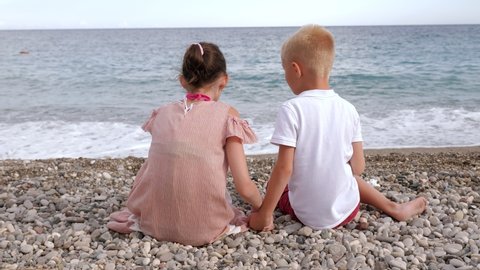 A little boy and a girl are sitting on the beach on a pebble and throwing stones into the water. A family with children on vacation by the sea.