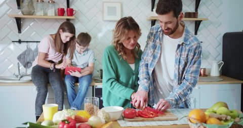 Young adorable family of four people resting in the kitchen. Funny flirting handsome married couple of parents cutting vegetable together cooking lunch having fun.