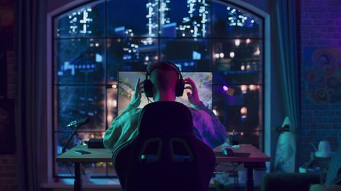 Back of the Head Angle of a Successful Stylish Gamer Putting on Headphones and Playing a Video Game on Personal Computer in a Neon Lit Living Room at Home. Young Man in Victorious.