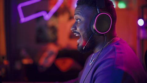 Gaming at Home: Gamer Playing Online Video Game on Computer. Close Up Portrait of Stylish Black Male in Headphones Enjoying Leisure Time, Talking with Players on Mic. Cyber Gaming Retro Neon Room.