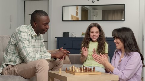 Happy family of three people of different races play chess at home in the living room. Mom wins dad. Game On, Family Meeting, Multi Ethnic Family, Different Generations.