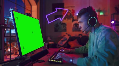 Excited Gamer Playing Online Video Game with a Mock Up Green Screen on His Powerful Personal Computer. Room and PC have Colorful Neon Led Lights. Cozy Evening at Home in Loft Apartment.