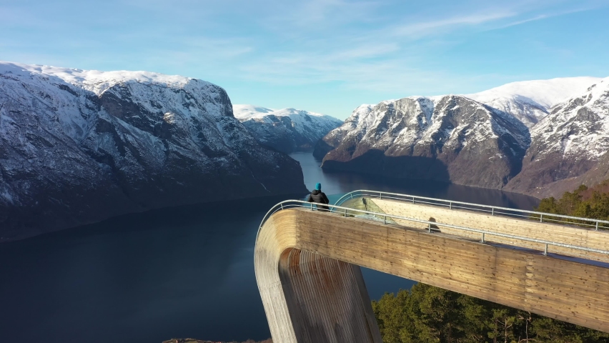 Flying slowly above man relaxing on edge of spectacular Stegastein viewpoint - Aurlandsfjorden and sognefjord in background - Norway winter aerial Royalty-Free Stock Footage #1089512901