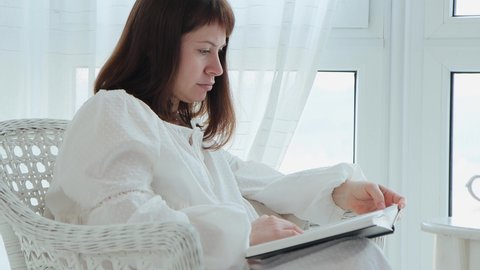 Profile portrait of a woman in white nightgown sits in armchair and reads a book thoughtfully at home, 4k video