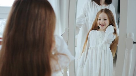Mom is combing yer cute red hair daughter and child girl is admiring yourself while spinning in front of the mirror, 4k portrait.