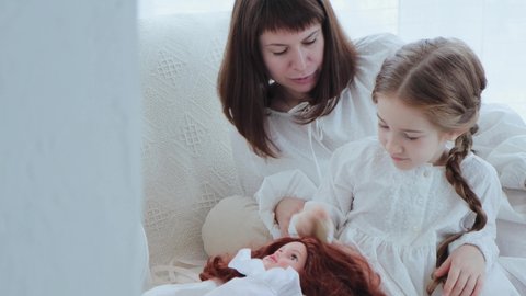 Mother with her little daughter sits on the sofa and girl plays with a doll. Child girl combs doll's hail and woman smile while looks on daughter. Panning from behind the wall, 4k video