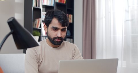 Close up of young Arab handsome man working at laptop and getting good news or great result in job. At home. Arabian joyful male freelancer at computer. Gamer winning in game online. Lockdown concept.