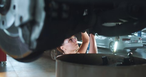 Young Caucasian woman in blue overalls, gloves and protective glasses tightens bolt under car using spanner. Female car mechanic at work in spacious garage.