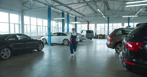 Young Caucasian woman in blue overalls and gloves holding spanner walks towards camera and smiles, while looking forward. Female car mechanic at workplace in spacious repair shop.