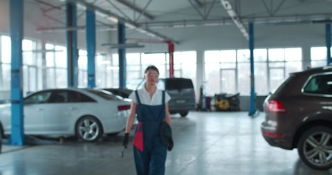 Young Caucasian woman in blue overalls, gloves and protective glasses walks towards camera holding spanner and smiles, while looking forward. Female car mechanic at workplace in spacious repair shop.