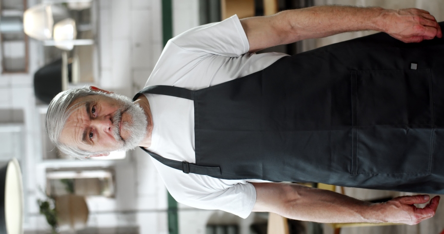 Bearded grey-haired man in apron talks to camera, gesturing with hands, giving instructions or explanations. Vertical shot of senior gentleman standing in cafe and speaking.