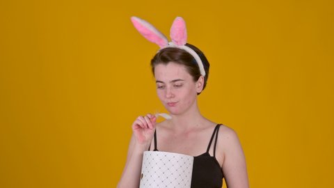 Young funny girl with bunny ears opens a gift box and is disappointed isolated on yellow background