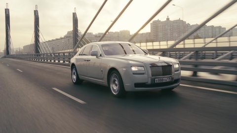 Saint-Petersburg, Russia - October, 2020: A white luxury car is driving over a bridge in the city at dawn. The camera shots in motion driving a Rolls-Royce Phantom.