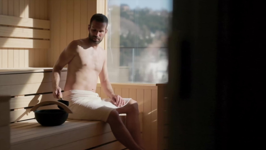 Handsome young man pouring water onto hot stone in the sauna