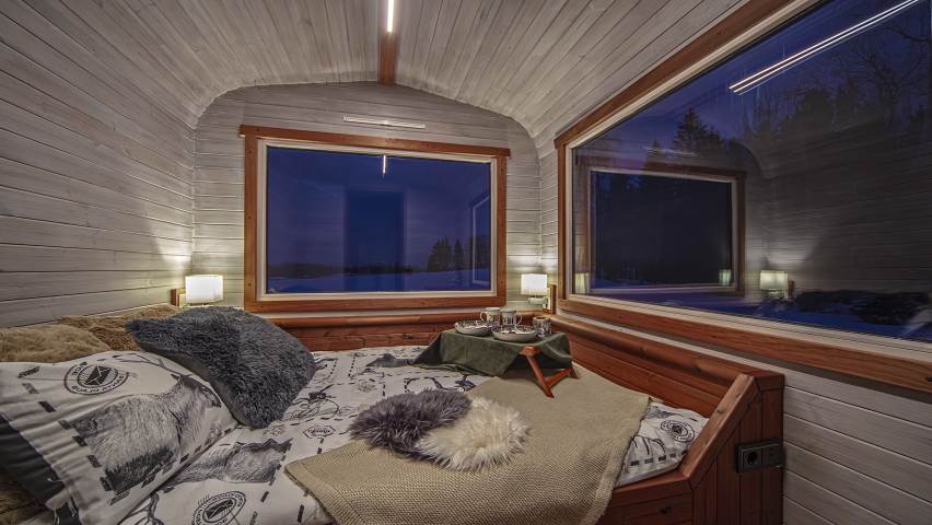 Static view of a cozy bedroom in a wooden cabin with huge glass window looking outside to the snow covered countryside at dawn in timelapse.