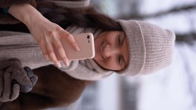 Charming brunette woman use mobile phone happy texting in social networks addicted to technologies, look at camera, smile, wear casual winter clothes on gray background. Vertical video