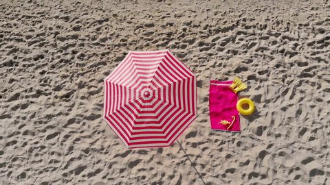 Striped sun protection umbrella near towel with rubber ring and snorkeling gear on sand. Sea waves wash shoreline on sunny day. Beach essentials in summer aerial view