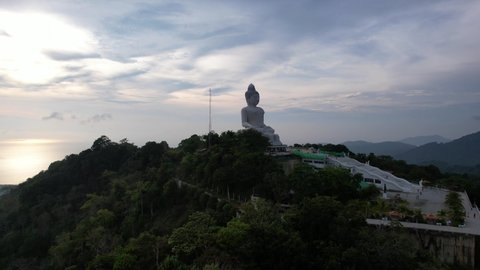 Drone view of the Big Buddha, Thailand. The Big Buddha is sitting on hill in the lotus position, meditating. The sun shines brightly through clouds. There's a jungle all around. A ray of sun on water