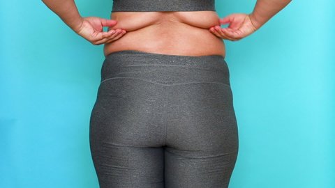 Back view of unrecognizable fat woman wearing grey leggings, sports bra, shaking, squeezing excess flabby back fat with hands on blue background. Body positive, obesity, weight loss, liposuction.