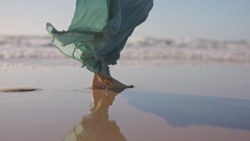 Slow motion shot of woman feet walk along ocean beach. Woman in long flowing dress walks along the ocean. UHD, 4K