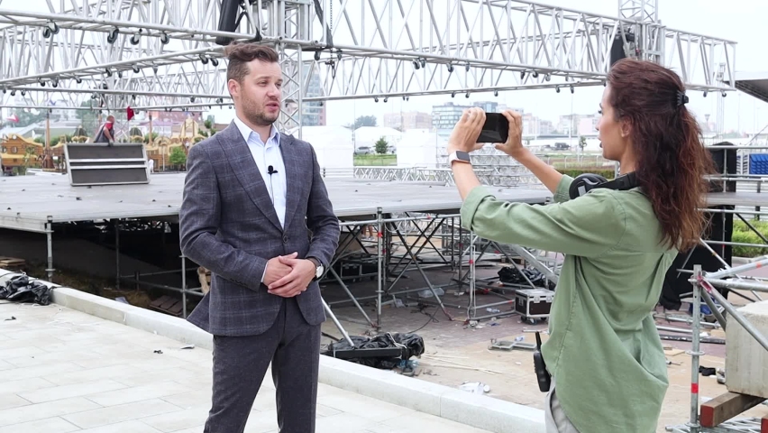 Assistant installs a microphone on the TV host. Preparing for interview. 
Blogger makes video.
Event manager report about stage construction. 
Installation of stage equipment. Live concert open air. 