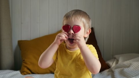 Child Boy with hearts on the eyes. Portrait of positive little kid close cover his eyes small red hearts. Toddler wear yellow trendy clothes on modern interior of domestic room. Cinematic footage.