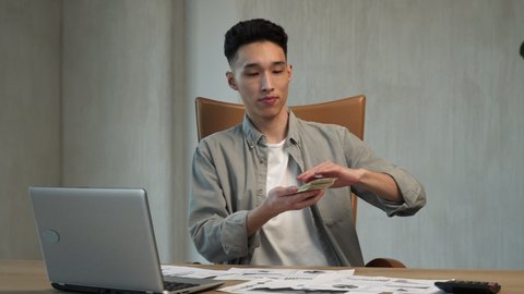 Happy Asian businessman throws dollar bills fanning out on wooden table after getting cash. Young man with money sits on chair by laptop at work in office