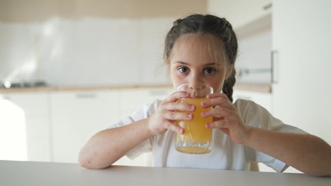 Little cute girl drinks fresh fruit juice. Happy child drinking orange drink. Healthy breakfast for a child. Girl drinks orange juice at the table in the kitchen. Healthy drink for a healthy lifestyle