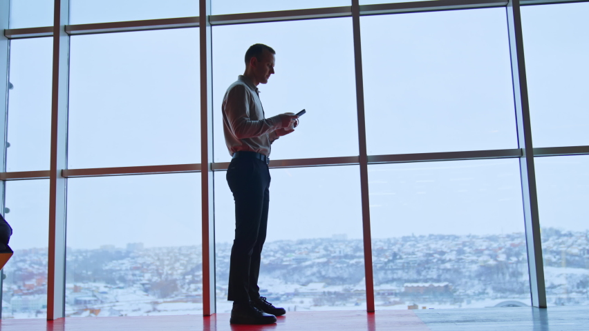 Young man standing tall at panoramic window with a cup and phone in hands. Full-length portrait of a male worker in office during lunch time.