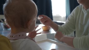Mother feeding her baby with spoon, one year old baby boy in high chair eating porridge. Morning sunlight from the window illuminates mother with child in the room. High quality 4k footage