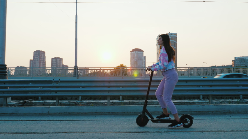 Beautiful girl riding electric kick scooter on sidewalk parallel to road in big city at sunset. Young woman travels in metropolis using e-scooter. Student moving on rental ecological transport Royalty-Free Stock Footage #1089548049