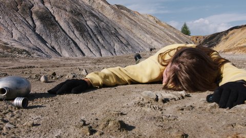 Girl exhausted from thirst lies in rocky wasteland with empty metal flask. Becoming weak hungry woman sprawled on ground. Drunk person is on sandy surface. Helplessness, early death in stone badlands