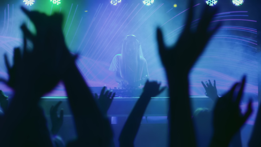 DJ plays disco music on turntable at night club scene. Partying joyful crowd moves in hip-hop rhythm of funky dance. Trendy girl in bright spot light of multi-color neon. Female dj at concert stage Royalty-Free Stock Footage #1089548495